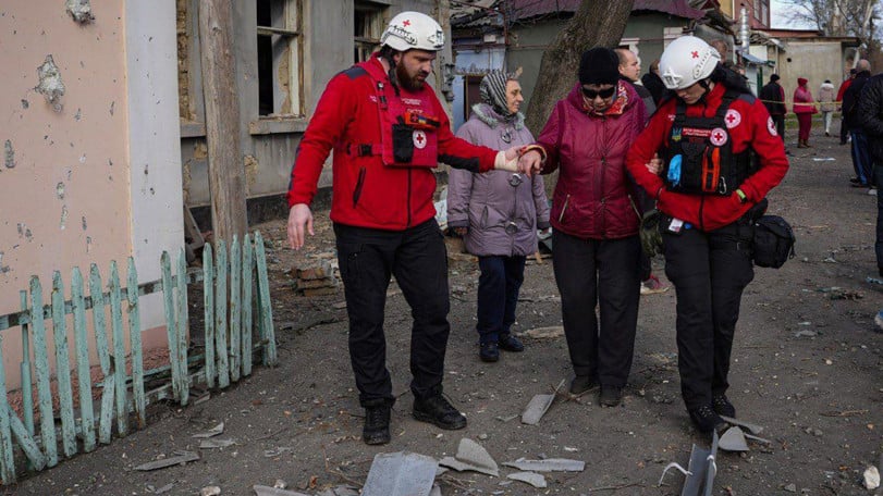 Två volontärer från Röda Korset hjälper en äldre kvinna att ta sig förbi bråte som ligger på marken i ett bostadsområde i staden Mykolajiv.