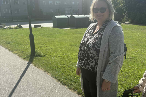 kvinna står utomhus med gräsmatta i bakgrunden, bredvid henne ser man en person vars händer och rullator syns.