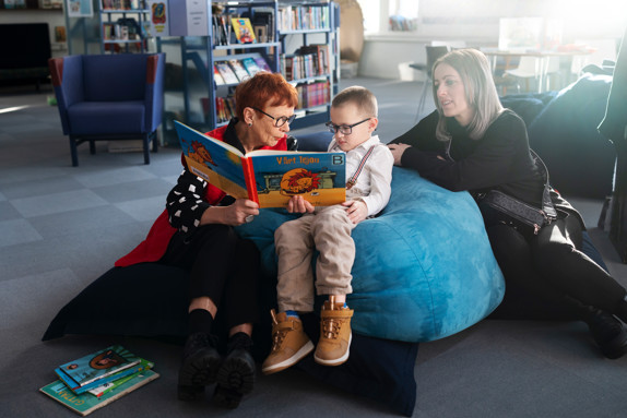 Volontären och läsfaddern Lena Olsson och Eman Begic sitter på kuddar och läser en bok. Bredvid sitter Emans mamma Alma.