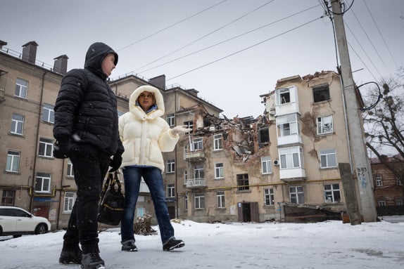 En man och en kvinna går med vinterjackor på en gata i Sumy. I bakgrunden syns ett hus som förstörts av kriget.