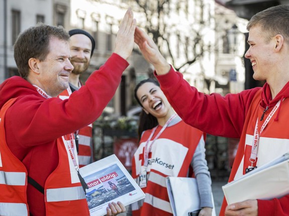 Två glada värvare i Svenska Röda Korset-kläder gör high five. I bakgrunden syns ytterligare två glada värvar-kollegor.