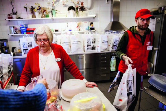 Två volontärer från Röda Korset står i Röda Korsets kök. Bullar står på en bänk och bakom volontärerna står matkassar på rad.