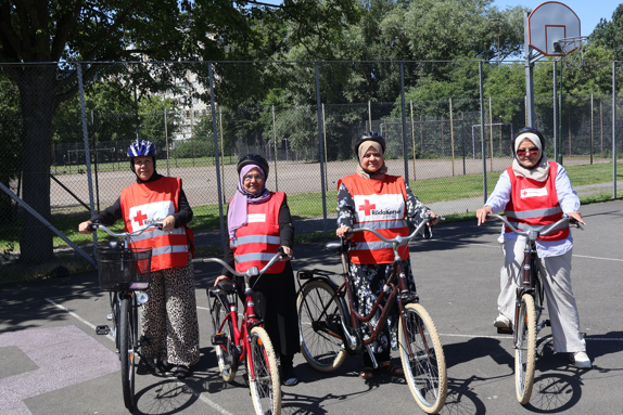 Fyra kvinnor som är volontärer i Röda Korsets cykelskola står med sina cyklar på en asfalterad uteplan i Lindängen, Malmö. De bär röda västar med Röda Korsets logotyp och cykelhjälm. I bakgrunden syns stängsel, träd och en basketkorg