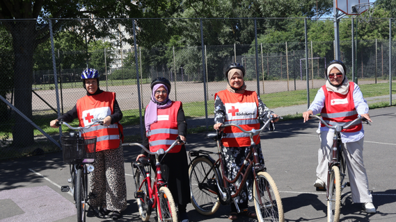 Fyra kvinnor som är volontärer i Röda Korsets cykelskola står med sina cyklar på en asfalterad uteplan i Lindängen, Malmö. De bär röda västar med Röda Korsets logotyp och cykelhjälm. I bakgrunden syns stängsel, träd och en basketkorg