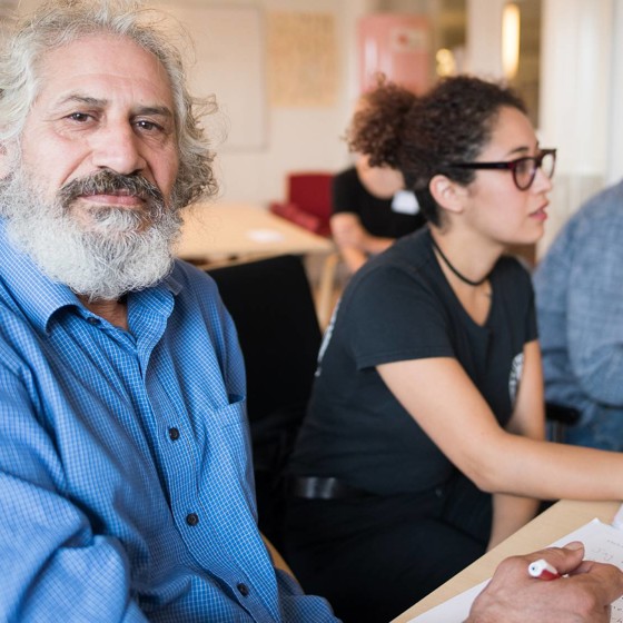 man i vitt skägg och blå skjorta sitter vid ett bord och tränar svenska