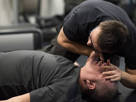 Bilden visar en man kollar ifall en annan man andas eller inte genom att hålla sin kind över munnen och titta ner på bröstkorgen.