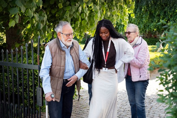 Volontär från Röda Korset promenerar utomhus med två äldre personer.
