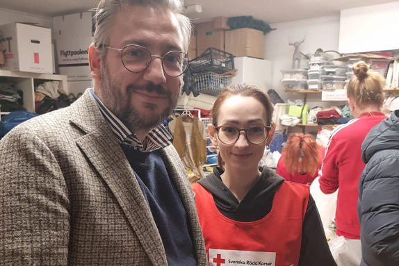 Foto på partiledaren Muharrem Demirok i kavaj och vår volontär Alex i en röd väst med Röda korsets-logotyp, i ett rum fullt av saker och lite människor i bakgrunden.