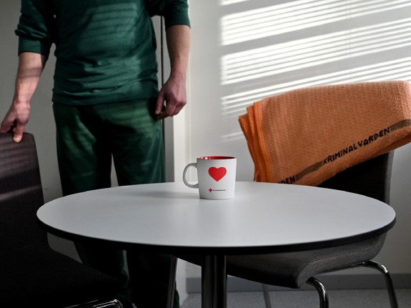 En man står vid ett bord och dragit ut en stol. Han är klädd i grön dräkt och på bordet står en kopp med ett hjärta på.