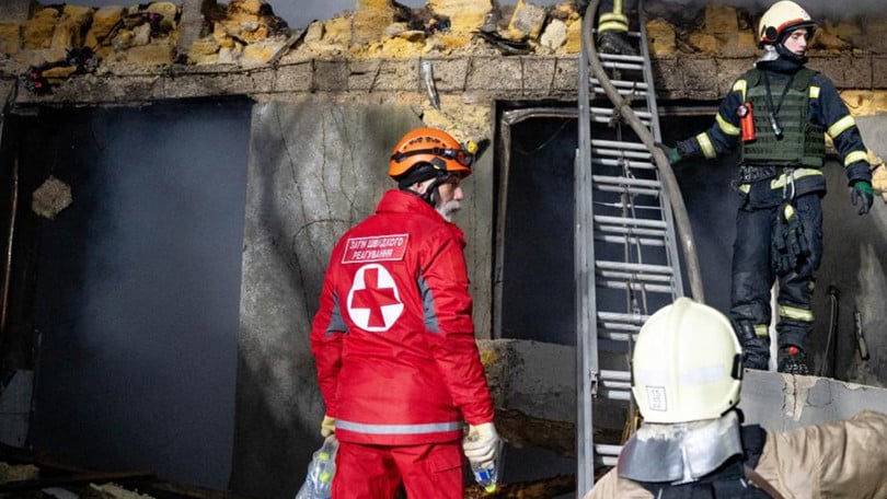 En Röda Korset volontär står jämte brandmän för att assistera i brandsläckningsarbetet framför en byggnad i ruiner som ryker.