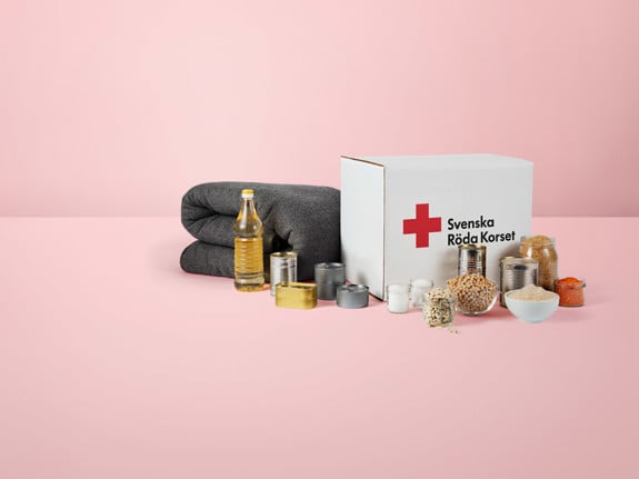 Ett matpaket som det står Svenska Röda Korset på. Uppställt framför paketet står dess innehåll i form av mat och konserver samt en grå ullfilt.