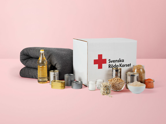 Ett matpaket som det står Svenska Röda Korset på. Uppställt framför paketet står dess innehåll i form av mat och konserver samt en grå ullfilt.