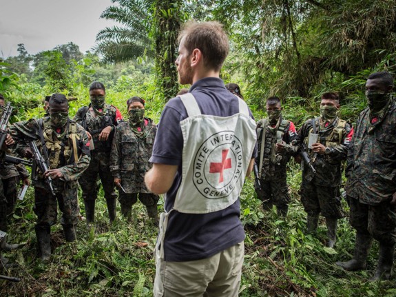 En volontär står inför soldater och pratar. Soldaterna är beväpnade med gevär och står i en halvcirkel runt volontären.