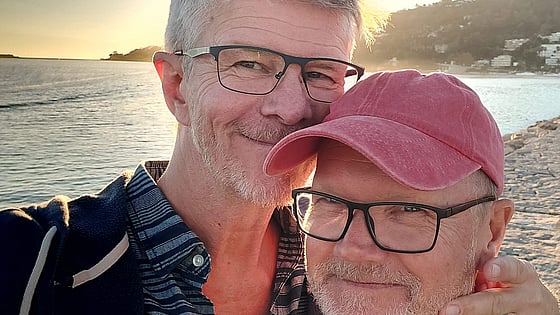 Två barn tittar fram bakom en dörr, som att de leker tittut. Det ena barnet visar hela ansiktet och ler in i kameran, det andra barnet syns bara ena ögat och lite av håret på.