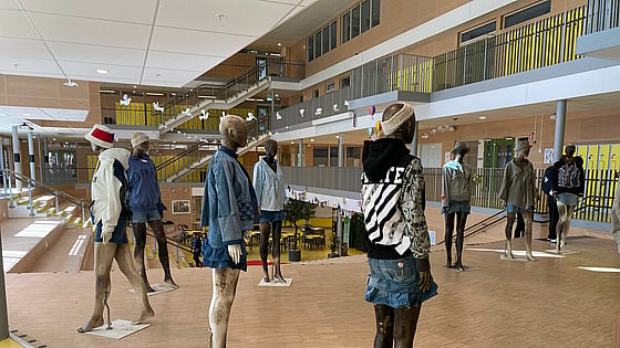 En rad skyltdockor står uppställda i en ljus skolmiljö. De bär kreativa outfits av återbrukade kläder i denim, hoodies och jackor med tryck. I bakgrunden syns öppna trappor, glaspartier och elever i rörelse – en miljö som förmedlar kreativitet, hållbarhet och lärande.