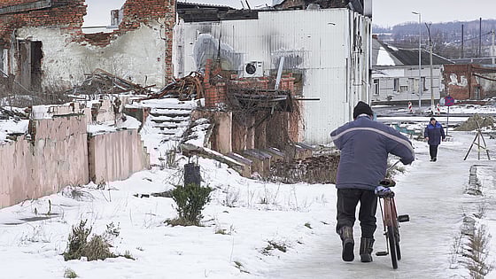 En man går med sin cykel på en gata i Sumy, det är snö på marken och mulet väder. Ett av husen är kraftigt skadat på grund av kriget.