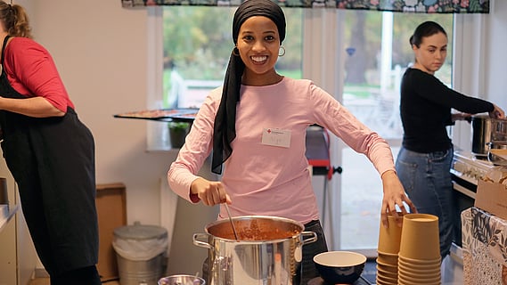 Bild från soppköket, kvinna i mitten av bilden tittar in i kameran och är i gång med att servera soppa. I bakgrunden syns två kvinnor till som håller på att förbereda mate och hjälper till med servering. 