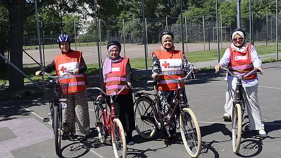 Fyra kvinnor som är volontärer i Röda Korsets cykelskola står med sina cyklar på en asfalterad uteplan i Lindängen, Malmö. De bär röda västar med Röda Korsets logotyp och cykelhjälm. I bakgrunden syns stängsel, träd och en basketkorg