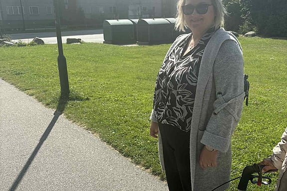 kvinna står utomhus med gräsmatta i bakgrunden, bredvid henne ser man en person vars händer och rullator syns. 