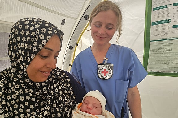Kvinna håller i sin bebis och ler. Bakom henne står vårdpersonal från Röda Korsets som också tittar på bebisen och ler.