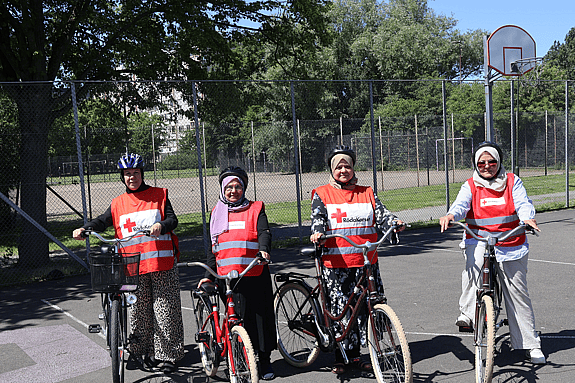 Fyra kvinnor som är volontärer i Röda Korsets cykelskola står med sina cyklar på en asfalterad uteplan i Lindängen, Malmö. De bär röda västar med Röda Korsets logotyp och cykelhjälm. I bakgrunden syns stängsel, träd och en basketkorg
