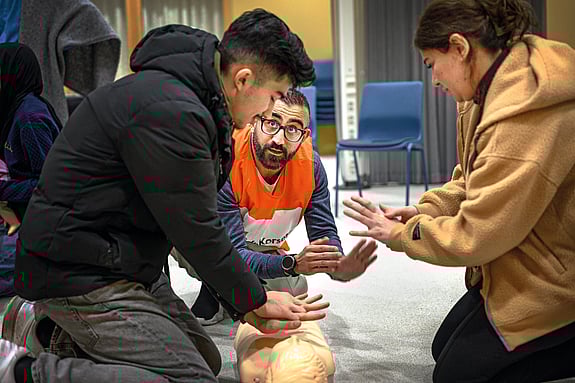 Volontär från Röda Korset instruerar en man och en kvinna i första hjälpen. De sitter på golvet och sträcker ut sina armar.