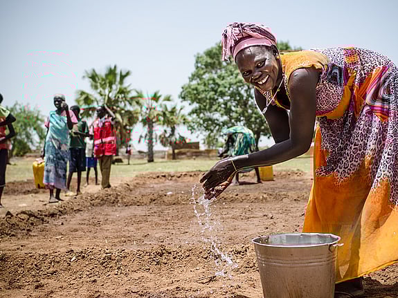 En kvinna tvättar händerna på ett fält. Hon ser glad ut och har färgglada kläder på sig.