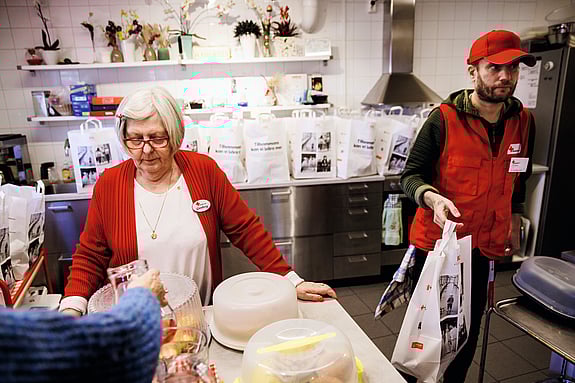 Två volontärer från Röda Korset står i Röda Korsets kök. Bullar står på en bänk och bakom volontärerna står matkassar på rad.
