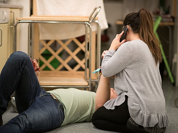 En kvinna sitter bredvid en man som ligger skadad på golvet och hon ringer samtidigt 112 med sin mobiltelefon.