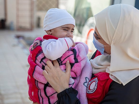 En syrisk röda korset-frivillig som springer genom rasmassor med en flicka i famnen. Det brinner i bakgrunden.