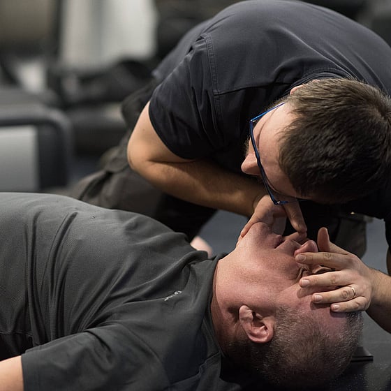 Bilden visar en man kollar ifall en annan man andas eller inte genom att hålla sin kind över munnen och titta ner på bröstkorgen.