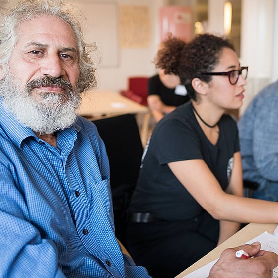 man i vitt skägg och blå skjorta sitter vid ett bord och tränar svenska