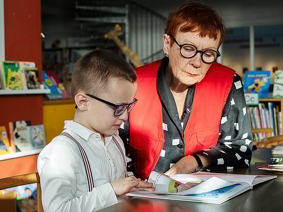 En pojke och en volontär sitter vid ett bord med en bok. Pojken har glasögon och läser ur boken medan volontären lyssnar.