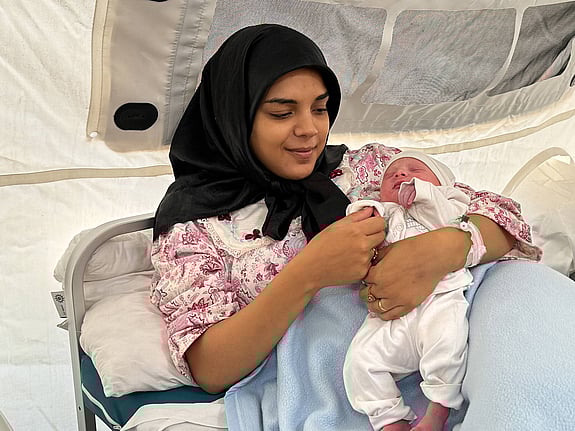 Kvinna ler och håller i sitt städbarn. Vårdpersonal från Röda Korset står bakom och tittar på barnet och ler.