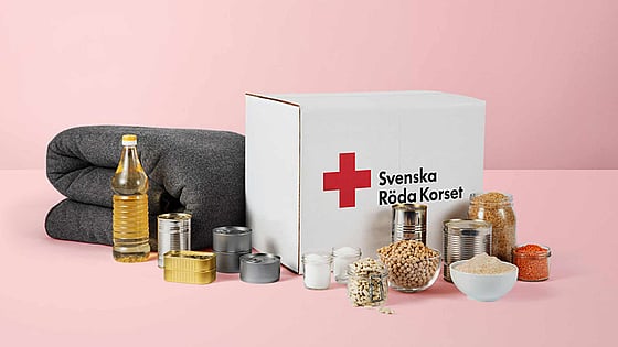 Ett matpaket som det står Svenska Röda Korset på. Uppställt framför paketet står dess innehåll i form av mat och konserver samt en grå ullfilt.