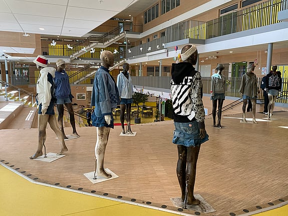 En rad skyltdockor står uppställda i en ljus skolmiljö. De bär kreativa outfits av återbrukade kläder i denim, hoodies och jackor med tryck. I bakgrunden syns öppna trappor, glaspartier och elever i rörelse – en miljö som förmedlar kreativitet, hållbarhet och lärande.