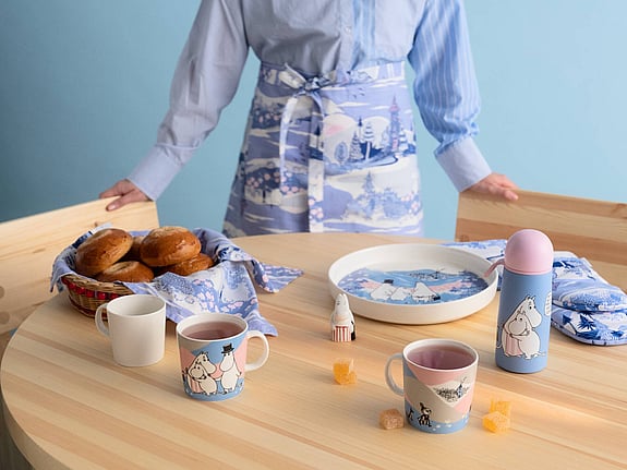 Porslin från Arabia med Mumin-figurer, står på ett träbord. I bakgrunden syns en person med förkläde.