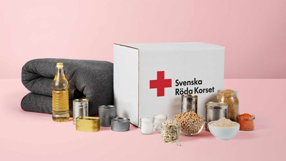 Ett matpaket som det står Svenska Röda Korset på. Uppställt framför paketet står dess innehåll i form av mat och konserver samt en grå ullfilt.