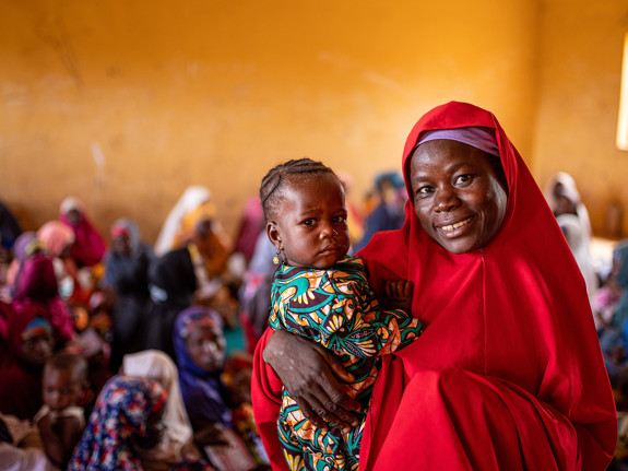 En mamma håller sitt barn i famnen. I bakgrunden syns andra människor.