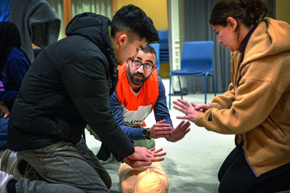 Volontär från Röda Korset instruerar en man och en kvinna i första hjälpen. De sitter på golvet och sträcker ut sina armar.