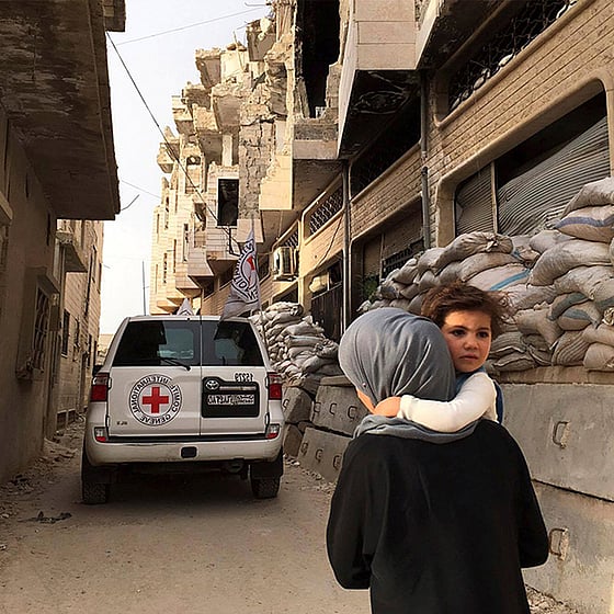 Barn bärs av sin mamma, i bakgrunden ser vi rasmassor och en röda kors-bil.