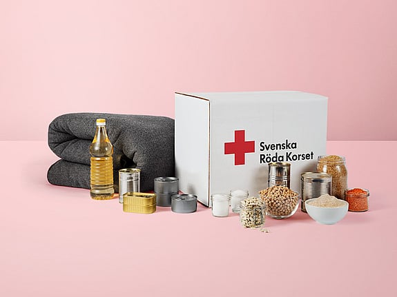 Ett matpaket som det står Svenska Röda Korset på. Uppställt framför paketet står dess innehåll i form av mat och konserver samt en grå ullfilt.